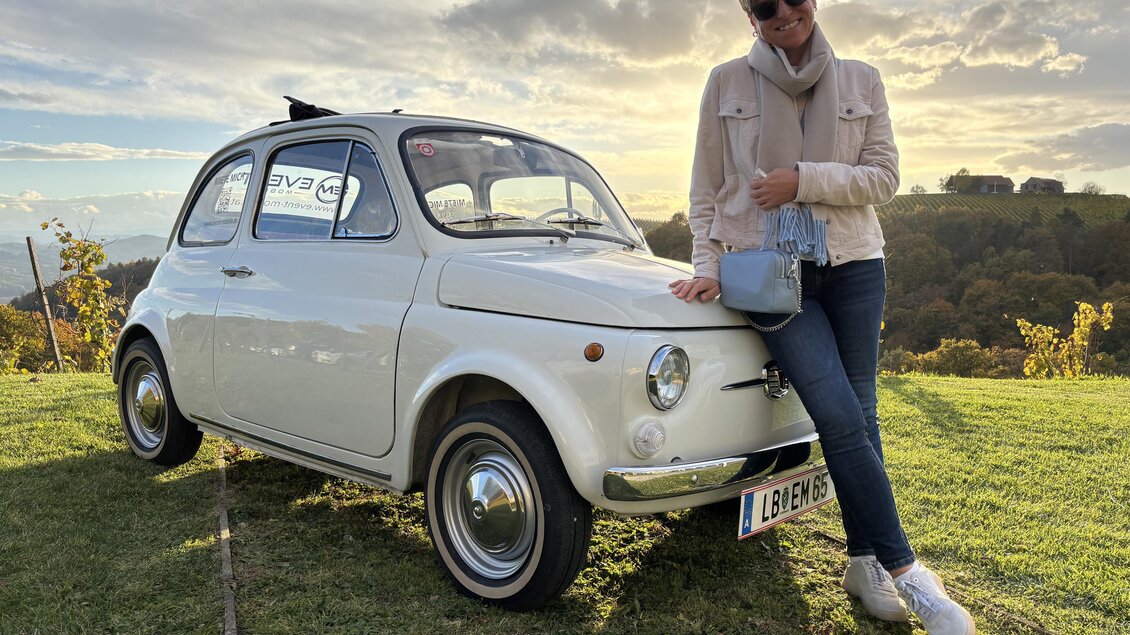 Eine Frau steht neben einem weißen Oldtimer-Auto auf einer Wiese. Im Hintergrund sind sanfte Hügel und ein bewölkter Himmel zu sehen.