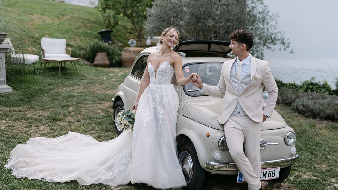 Ein Brautpaar steht neben einem kleinen Auto auf einer Wiese. Der Bräutigam hält die Hand der Braut, die ein wunderschönes Hochzeitskleid trägt.