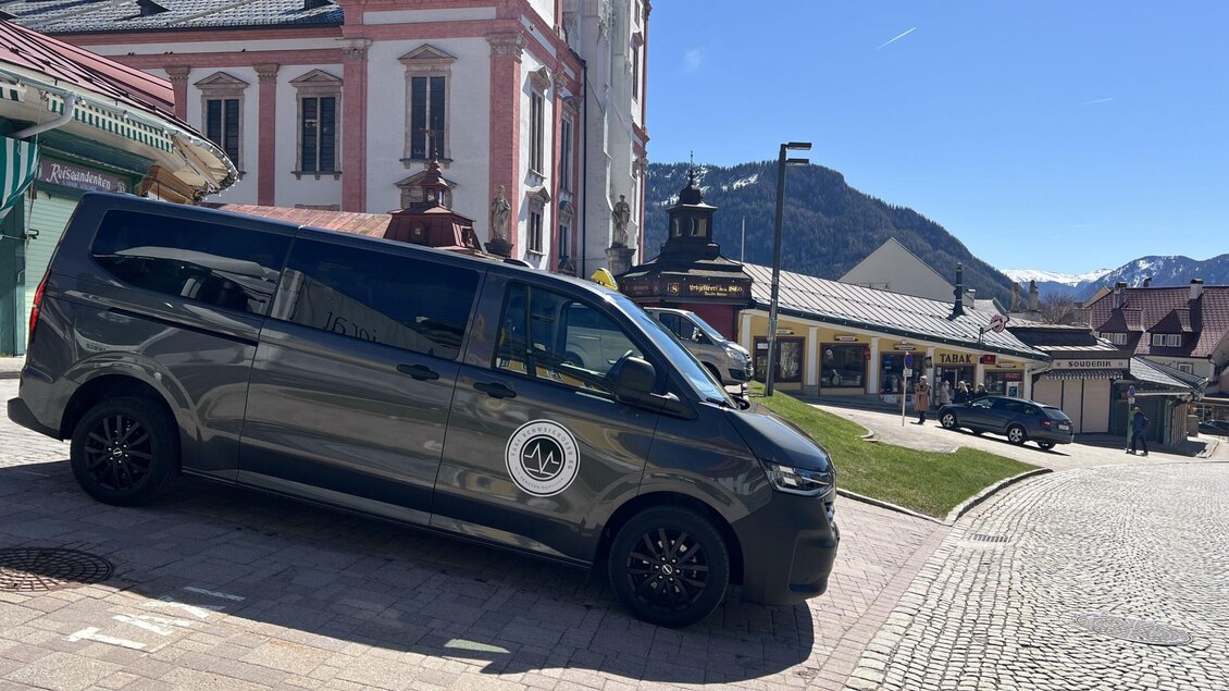 Ein grauer Van steht auf einem gepflasterten Platz in einer malerischen Stadt. Im Hintergrund sind historische Gebäude und Berge zu sehen. | © Taxi Schweighofer KG
