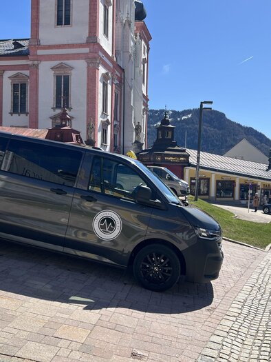 Ein grauer Van steht auf einem gepflasterten Platz in einer malerischen Stadt. Im Hintergrund sind historische Gebäude und Berge zu sehen. | © Taxi Schweighofer KG