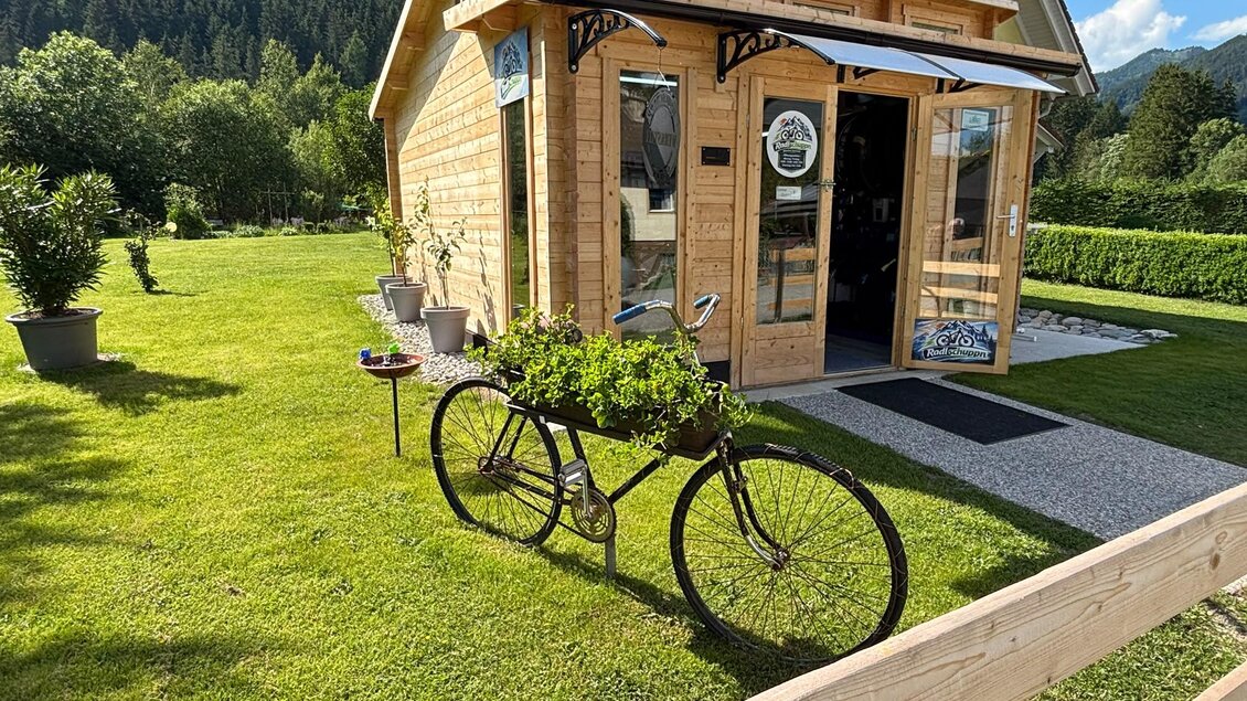 Ein modernes Holzhaus mit großen Fenstern steht in einer grünen Gartenlandschaft. Vor dem Haus steht ein Fahrrad mit einem Blumenkasten darauf. | © TV Hochsteiermark/Andrea Stelzer