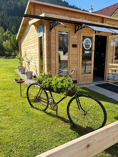 Ein modernes Holzhaus mit großen Fenstern steht in einer grünen Gartenlandschaft. Vor dem Haus steht ein Fahrrad mit einem Blumenkasten darauf. | © TV Hochsteiermark/Andrea Stelzer