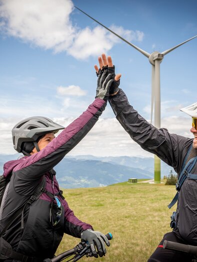 Zwei Radfahrer geben sich ein High Five auf einem Berghang. Im Hintergrund sind Windräder und eine prachtvolle Landschaft zu sehen. | © Jesse Streibl