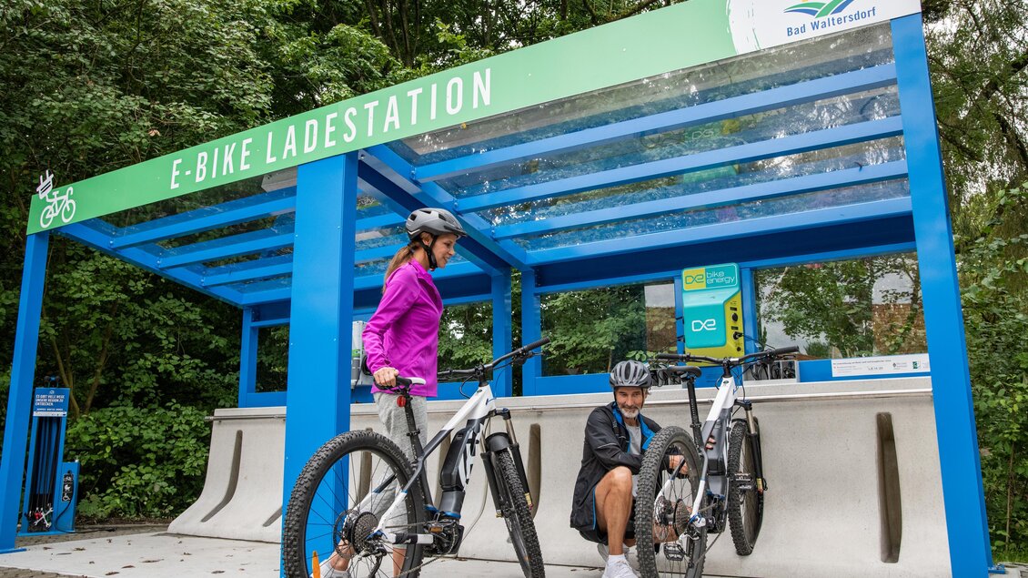 Eine E-Bike Ladestation mit zwei Fahrrädern. Zwei Personen stehen dabei und nutzen die Station. | © Thermen- & Vulkanland