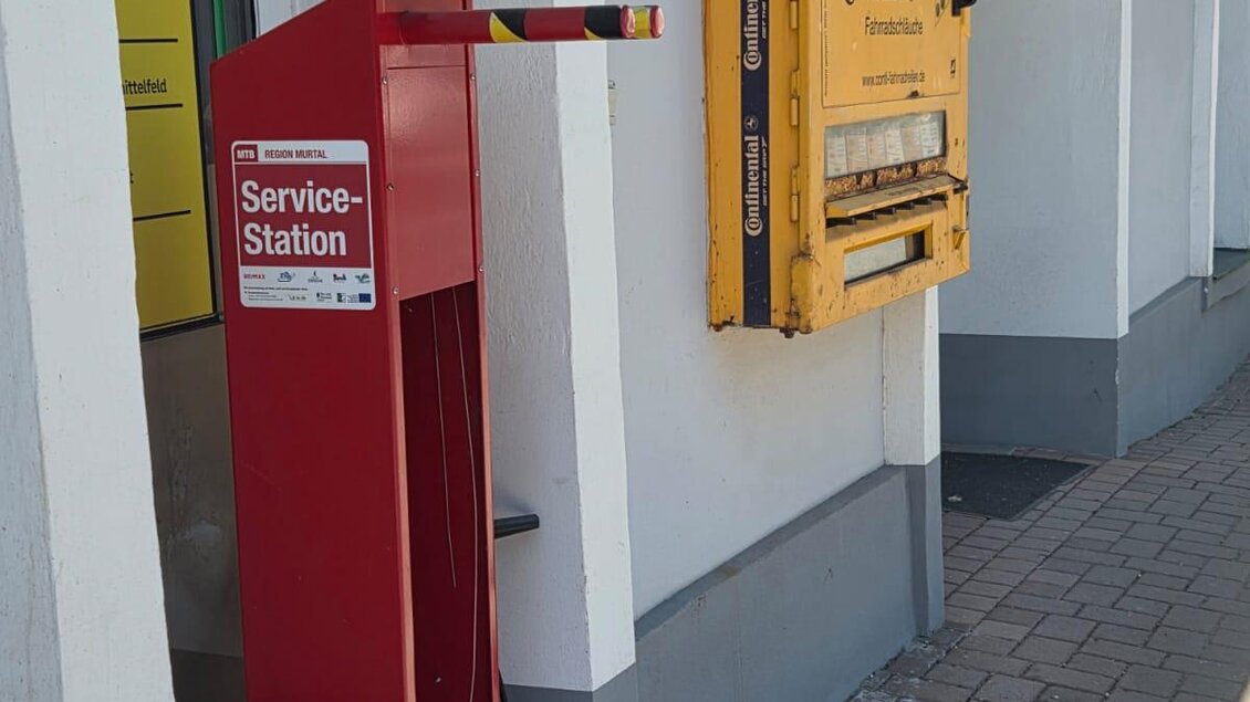 Eine rote Service-Station steht an der Seite des Weges. Im Hintergrund ist ein Schild mit der Aufschrift "Sportzen" zu sehen. | © Roman Gruber