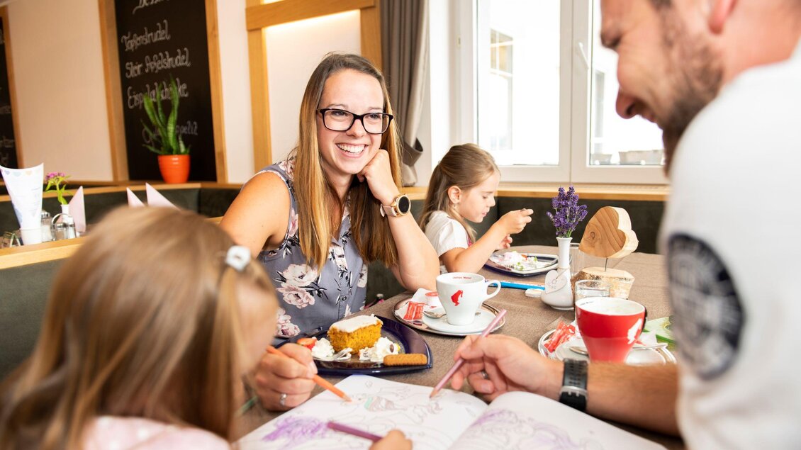 Eine fröhliche Familie sitzt in einem Café und genießt ihre Zeit zusammen. Die Kinder malen in einem Malbuch, während die Erwachsenen sich unterhalten. | © Familienhotel Berger