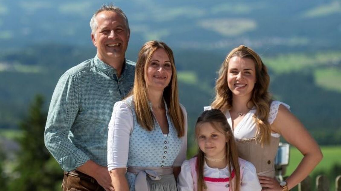 Eine fröhliche Familie in traditioneller Tracht steht auf einer Holzveranda. Im Hintergrund sind grüne Wiesen und sanfte Hügel zu sehen. | © Familienhotel Berger