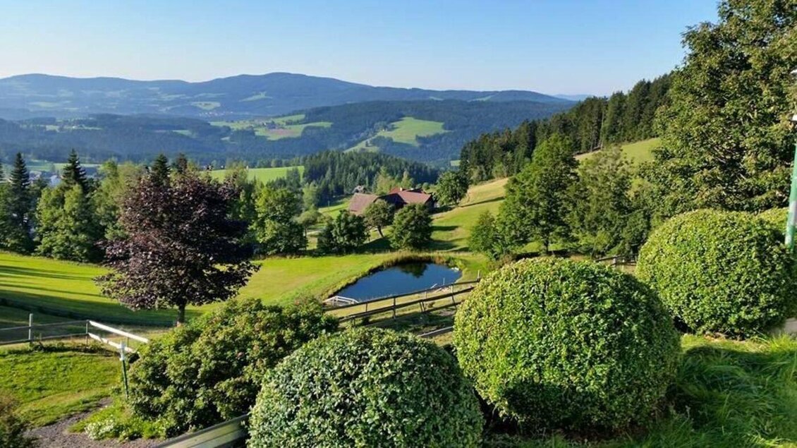 Eine malerische Landschaft mit sanften Hügeln, Bäumen und einem ruhigen Teich. Im Hintergrund sind Berge und eine klare blaue Skyline zu sehen. | © Familienhotel Berger