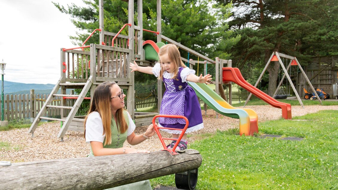 Eine Mutter spielt mit ihrer Tochter auf einem Spielplatz. Im Hintergrund sind Rutschen und ein Spielturm zu sehen. | © Familienhotel Berger