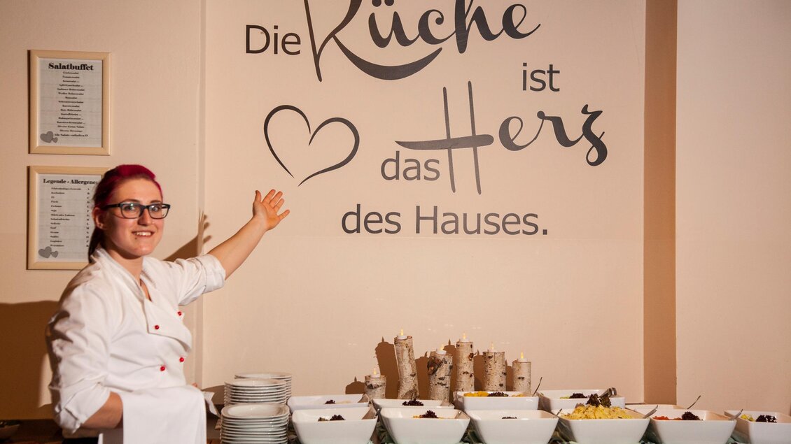 Eine Köchin steht vor einer Wand mit dem Spruch "Die Küche ist das Herz des Hauses." Auf einem Tisch sind verschiedene Speisen angerichtet. | © Familienhotel Berger