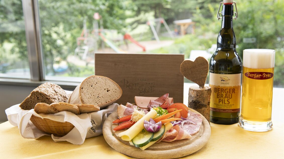 Ein Tisch mit verschiedenen Brotsorten, Wurst und Gemüse auf einem Holzbrett. Dazu steht eine Flasche Bier und ein Glas Bier. | © Familienhotel Berger