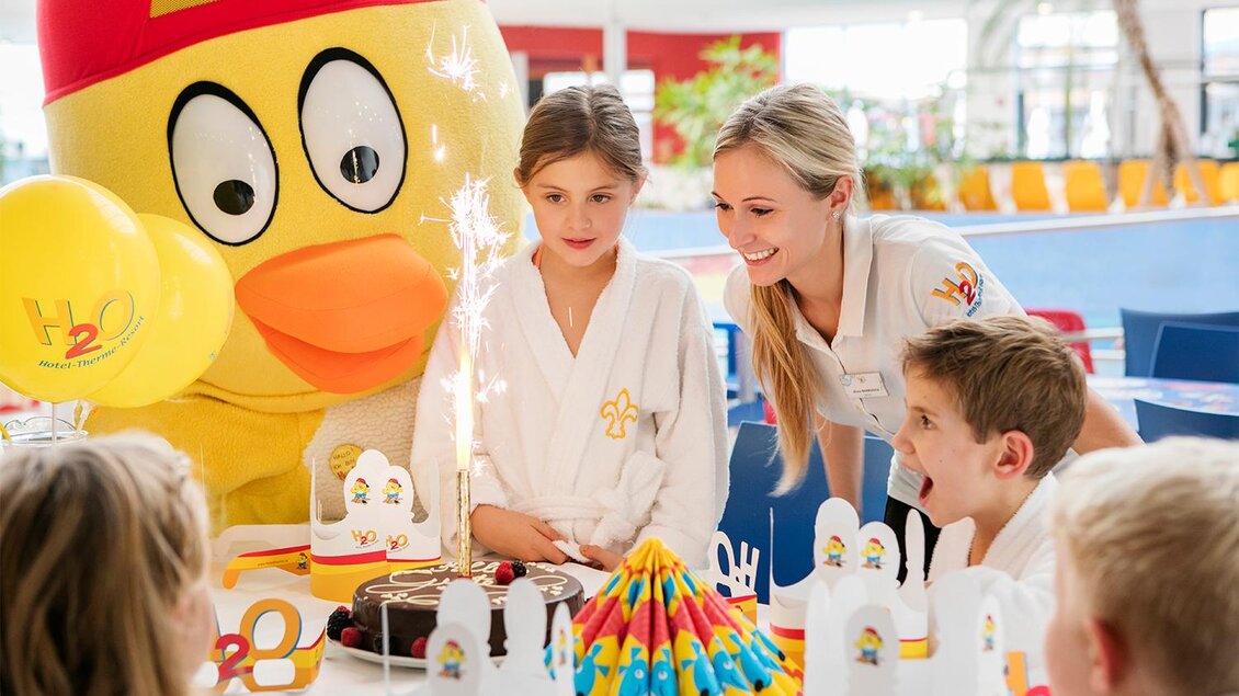 Ein Kindergeburtstagsfeier mit einem großen, gelben Entenmaskottchen. Kinder in Bademänteln sitzen um einen Tisch mit einer Torte und Partyhüten. | © Eisenberger