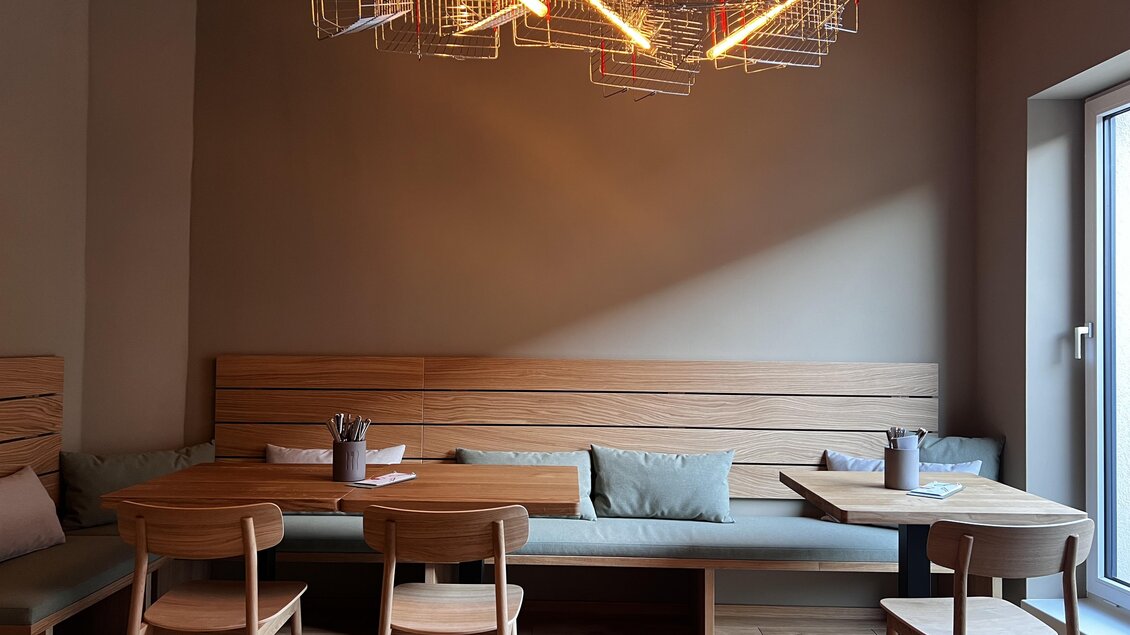 Ein modernes Café mit minimalistischer Einrichtung. Es gibt Holztische und -stühle sowie eine auffällige Lampe an der Decke. | © Feld Feldbach – Ein Marktlokal