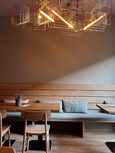 Ein modernes Café mit minimalistischer Einrichtung. Es gibt Holztische und -stühle sowie eine auffällige Lampe an der Decke. | © Feld Feldbach – Ein Marktlokal