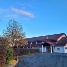 Ein modernes Gebäude mit Satteldach und einem Eingangsbereich. Es steht in einer ruhigen Umgebung mit Bäumen und einem blauen Himmel. | © TV Südsteiermark - Irene Löschnig