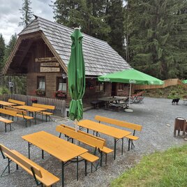 Eine gemütliche Berghütte umgeben von hohen Bäumen. Auf der Terrasse stehen mehrere Holztische mit grünen Sonnenschirmen. | © TVB Murau