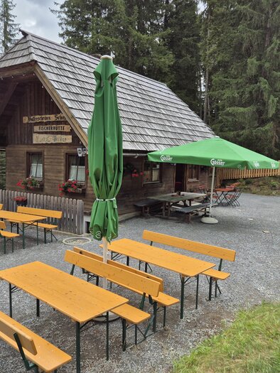 Eine gemütliche Berghütte umgeben von hohen Bäumen. Auf der Terrasse stehen mehrere Holztische mit grünen Sonnenschirmen. | © TVB Murau
