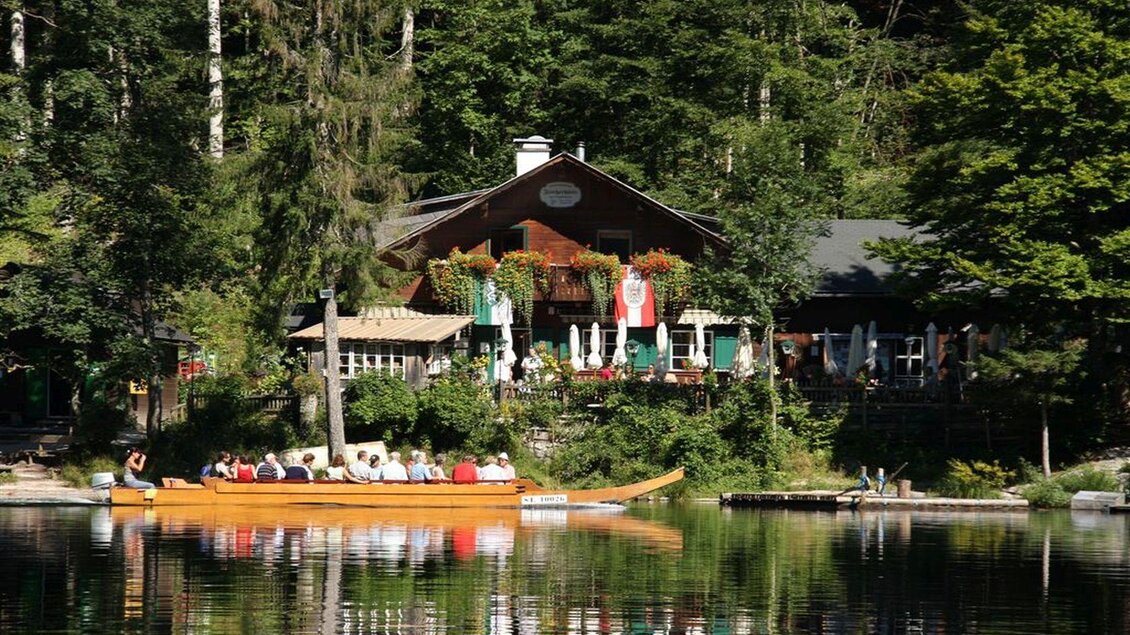 Ein malerisches Café am See, umgeben von grünen Bäumen. Im Vordergrund fährt ein Boot mit Passagieren über das ruhige Wasser. | © Albrecht Syen
