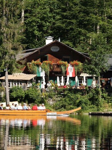 Ein malerisches Café am See, umgeben von grünen Bäumen. Im Vordergrund fährt ein Boot mit Passagieren über das ruhige Wasser. | © Albrecht Syen