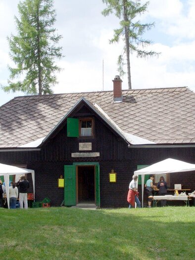 Ein traditionelles schwarzes Holzhaus mit grünen Fensterläden. Vor dem Haus stehen Zelte und Menschen, die an einem Event teilnehmen. | © Naturfreunde