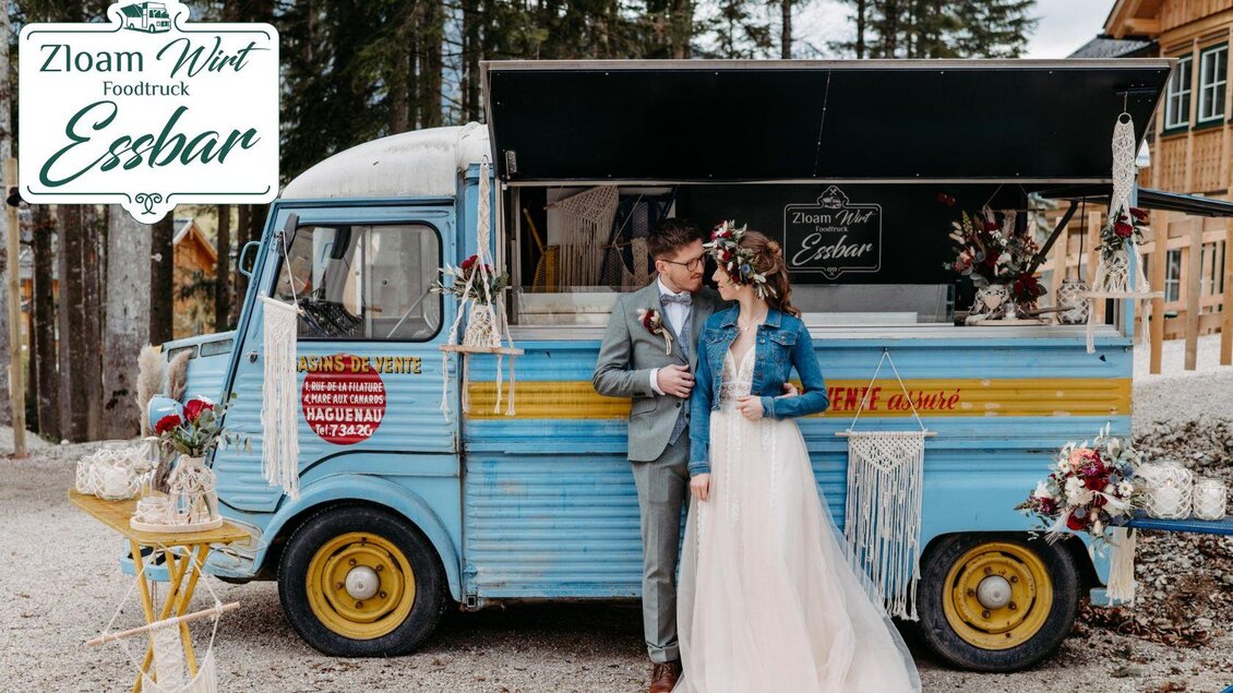 Ein Brautpaar steht vor einem bunten Foodtruck, der mit Blumen geschmückt ist. Der Truck trägt die Aufschrift „Zloam Wirt Essbar“ und befindet sich in einer naturnahen Umgebung. | © www.zloamwirt.at