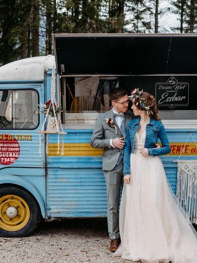 A bride and groom stand in front of a colorful food truck adorned with flowers. The truck bears the inscription "Zloam Wirt Essbar" and is situated in a natural setting. | © www.zloamwirt.at