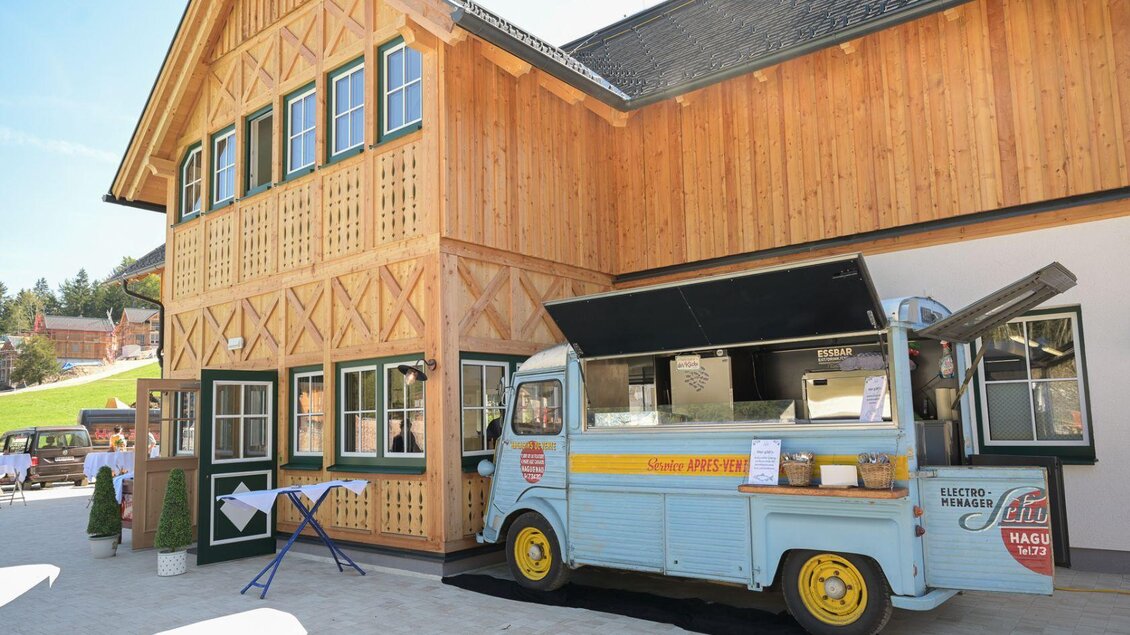 Ein hübscher Holzbau im rustikalen Stil neben einem altmodischen Essenswagen. Der Wagen bietet einladende Speisen in einer schönen Umgebung. | © Narzissendorf Zloam www.zloam.at