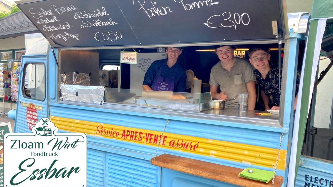 Ein bunter Foodtruck mit drei freundlichen Verkäufern, die Gerichte anbieten. Die Preisliste zeigt eine Auswahl an Speisen, darunter Pommes für €3,00. | © www.zloamwirt.at