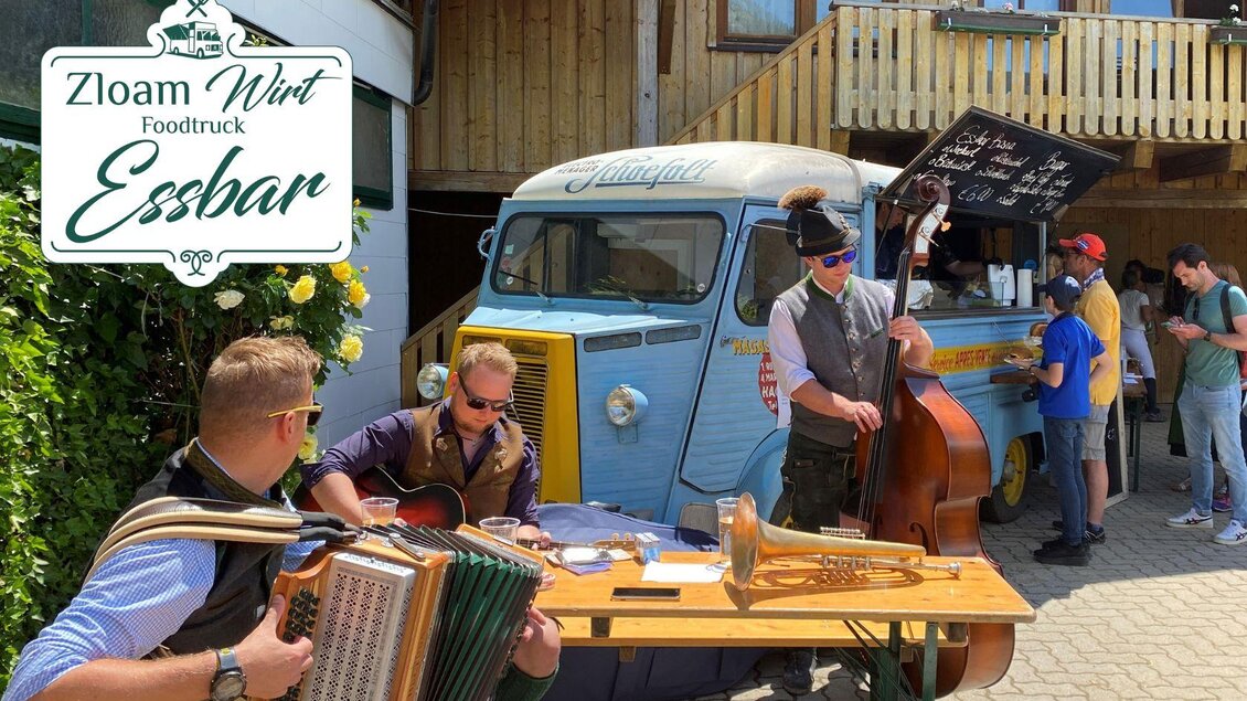 Ein Foodtruck mit der Aufschrift "Zloam Wirt Essbar" steht in einer sonnigen Umgebung. Musiker spielen traditionell, während Gäste im Hintergrund entspannen. | © www.zloamwirt.at