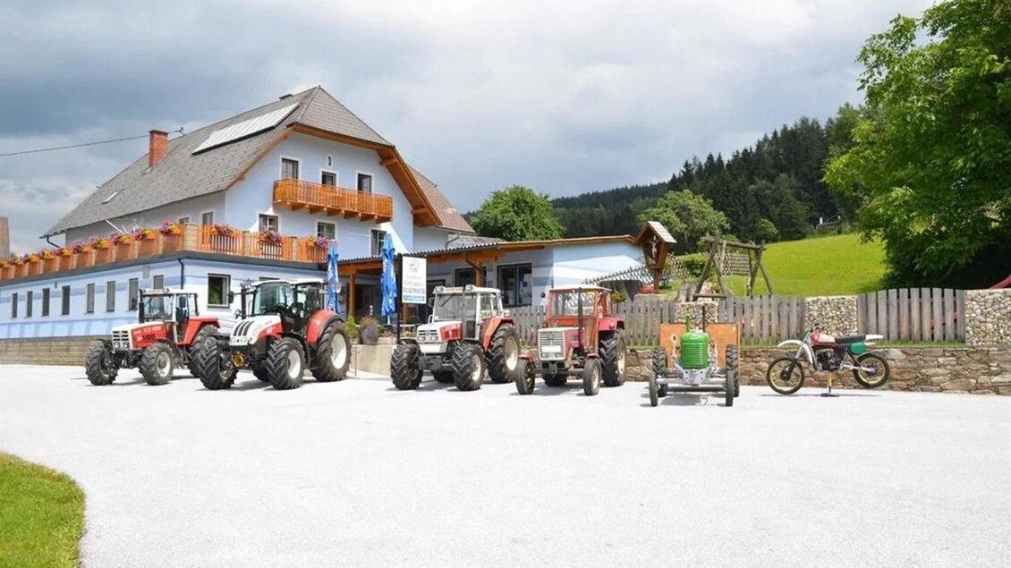 Ein Bauernhof mit einem großen Gebäude und mehreren Traktoren davor. Die Umgebung ist grün und hügelig, ideal für die Landwirtschaft. | © Forellengasthof Falkenbauer