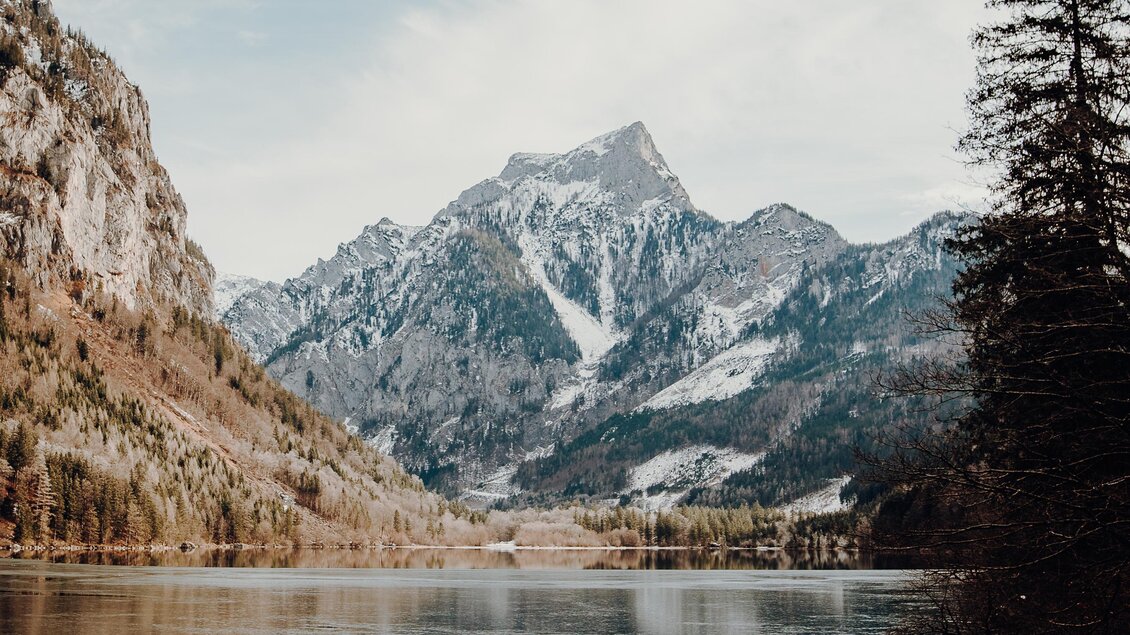 Ein ruhiger See umgeben von majestätischen Bergen. Der Himmel ist leicht bewölkt und reflektiert sich im Wasser. | © GenussReich