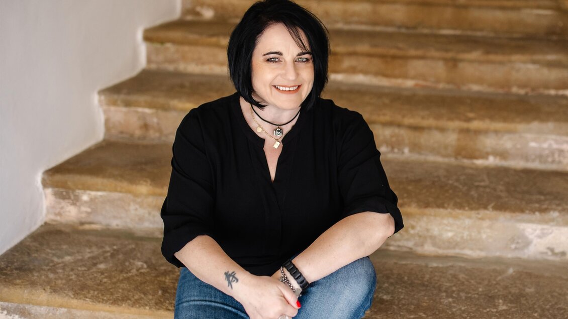Eine Frau sitzt auf einer Treppe und lächelt freundlich. Sie trägt ein schwarzes Oberteil und Jeans. | © Karin Bergmann