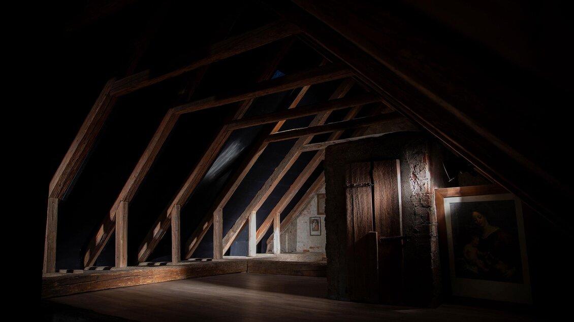 Ein düsterer Dachboden mit schrägen Holzbalken und Fenstern. Es gibt einen alten Schrank und ein Bild an der Wand. | © Nicole Oberhofer – FotoGrafin