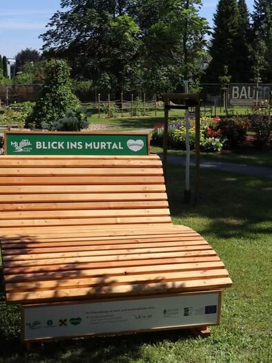 Eine Holzliege im Park mit der Aufschrift "BLICK INS MURTAL". Im Hintergrund sind Bäume und eine gepflegte Gartenlandschaft zu sehen. | © Erlebnisregion Murtal