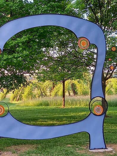 Eine moderne Skulptur in einem grünen Park. Sie hat eine geschwungene Form und farbige Kreise, die an eine Zielscheibe erinnern. | © Thermen- & Vulkanland
