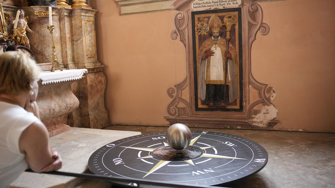 Eine Kompassplatte mit einer glänzenden Kugel, die sich darauf bewegt. Im Hintergrund ist ein historisches Wandbild sichtbar. | © Tourismusverband Oststeiermark