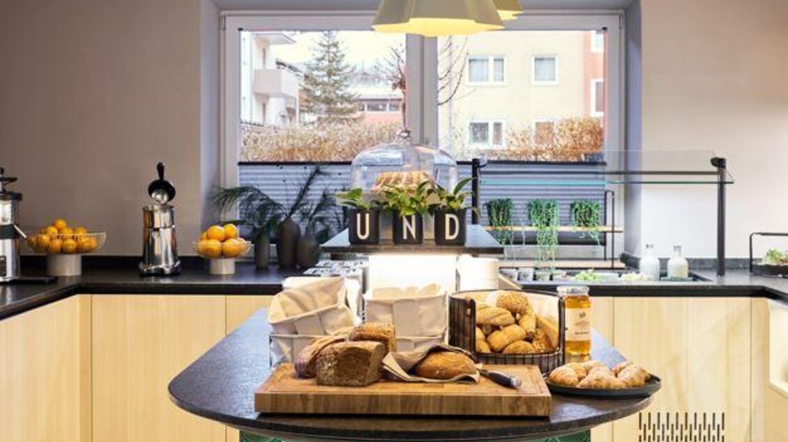 Eine moderne Küche mit einem großen Tisch in der Mitte. Auf dem Tisch befinden sich verschiedene Backwaren und Obst. | © @hotelund.me 