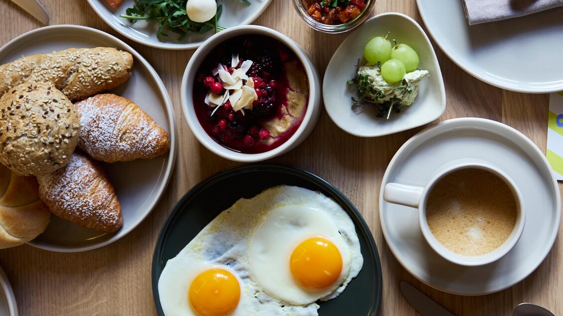 Ein reichhaltiges Frühstücksarrangement mit Eiern, frischem Obst, Joghurt und Gebäck. Kaffee und kleine Schalen mit verschiedenen Beilagen ergänzen das Bild. | © @hotelund.me 
