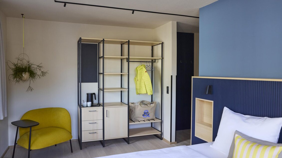 Ein modernes Zimmer mit einem bequemen gelben Stuhl und einem offenen Regal. Die Wände sind in hellen Farbtönen gestrichen, und es gibt eine gemütliche Bettgarnitur mit einem gestreiften Kissen. | © @hotelund.me 