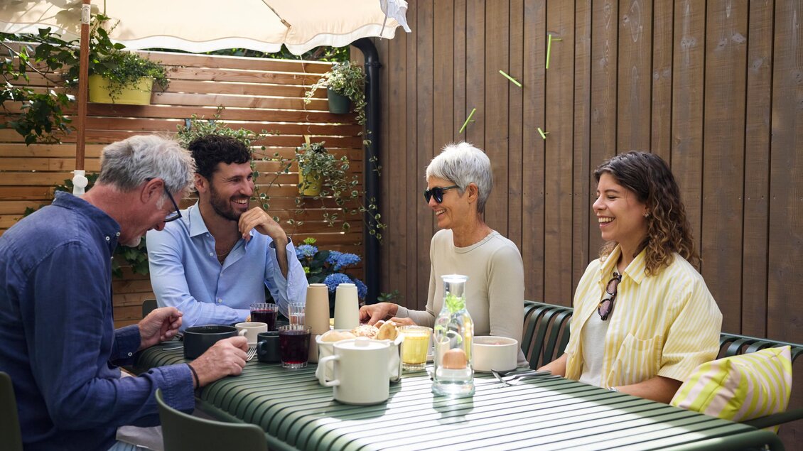 Eine Gruppe von vier Personen sitzt an einem Tisch im Freien. Sie lachen und unterhalten sich in einer gemütlichen Atmosphäre.