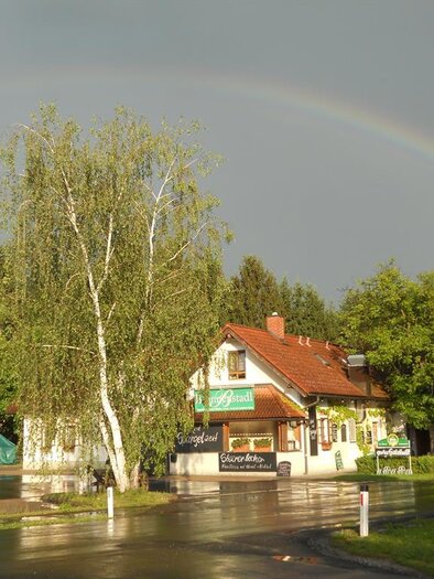 Ein ruhiges Straßenbild mit einem kleinen Gebäude und grünen Bäumen. Ein Regenbogen spannt sich über den Himmel nach einem Regenschauer.