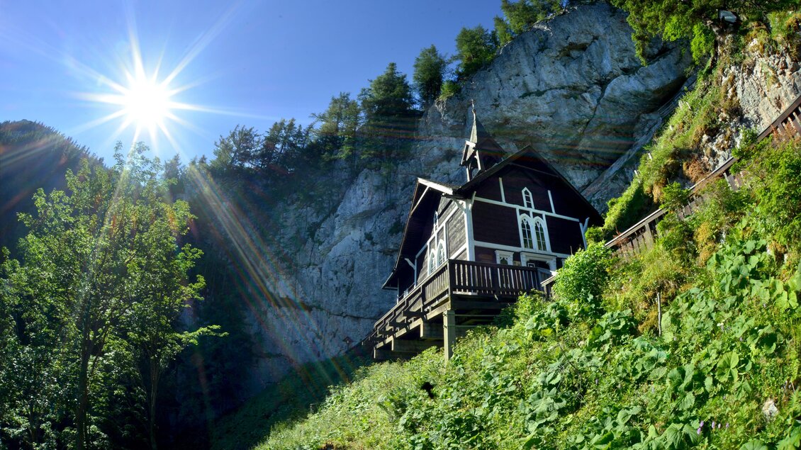 Ein malerisches Haus am Fuße einer Felswand, umgeben von üppigem Grün. Die Sonne scheint hell über die Landschaft. | © Tourismusverband Oststeiermark