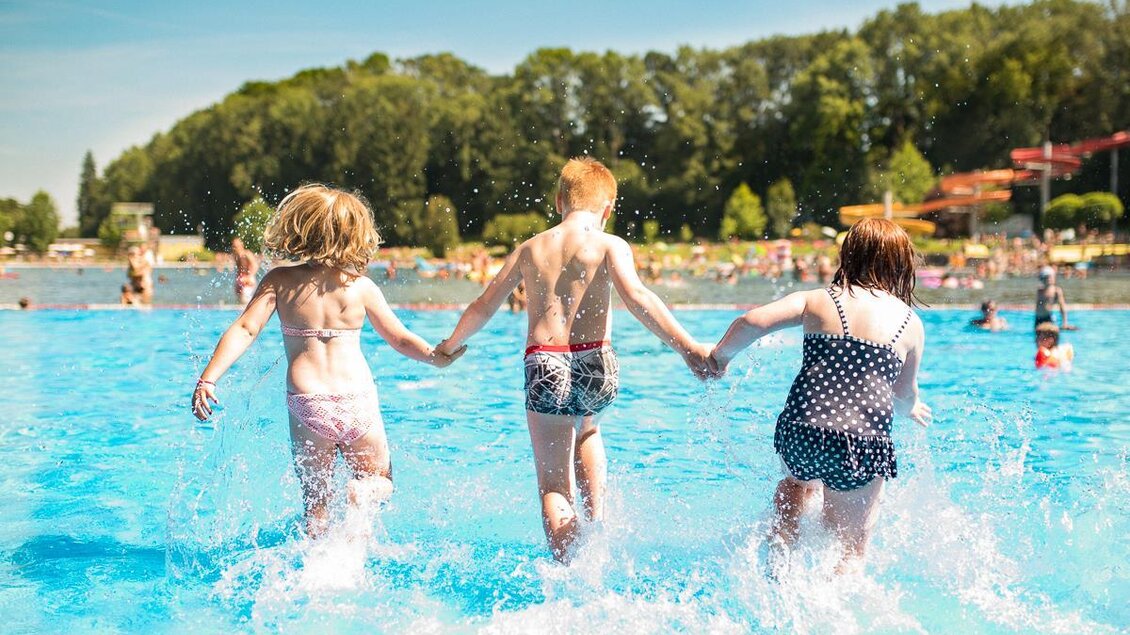 Drei Kinder laufen fröhlich ins Wasser und halten sich an den Händen. Im Hintergrund sind weitere Badegäste und eine grüne Landschaft zu sehen. | © Tourismusverband Thermen- & Vulkanland