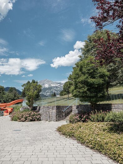 Eine ruhige Landschaft mit einem gepflasterten Weg, umgeben von Bäumen und bunten Blumen. Im Hintergrund sind Berge und ein klarer blauer Himmel zu sehen. | © TV Gesäuse