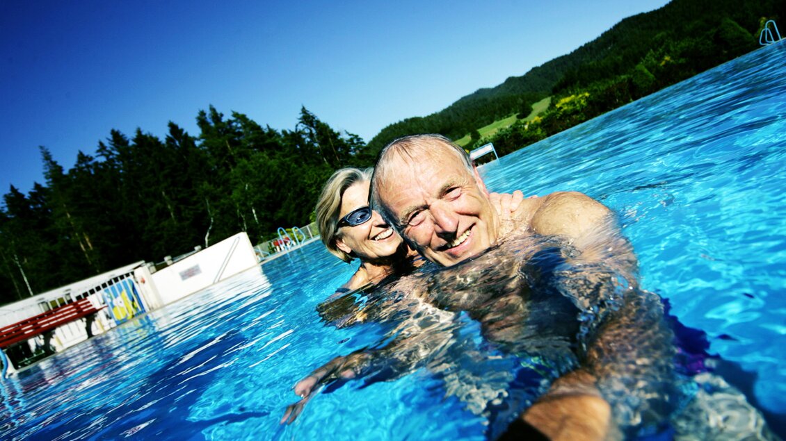 Ein fröhliches Paar schwimmt im Pool unter klarem, blauem Himmel. Die Umgebung ist grün und sonnig, perfekt für Entspannung. | © weinfranz.at
