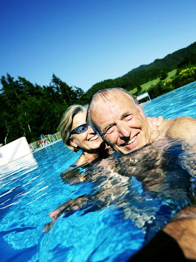 Ein fröhliches Paar schwimmt im Pool unter klarem, blauem Himmel. Die Umgebung ist grün und sonnig, perfekt für Entspannung. | © weinfranz.at
