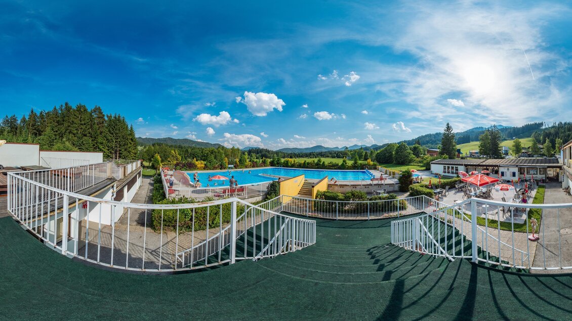 Ein Blick auf einen großen Poolbereich mit Liegen und Sonnenschirmen. Im Hintergrund sieht man grüne Berge und einen strahlend blauen Himmel. | © TV Mitterbach