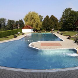 Ein großer Swimmingpool mit klarem Wasser und einer Wasserrutsche. Umgeben von einer grünen Wiese und Bäumen gibt es Sitzgelegenheiten und ein Gebäude im Hintergrund. | © Stadtgemeinde Fürstenfeld