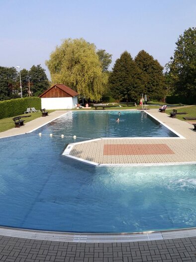 Ein großer Swimmingpool mit klarem Wasser und einer Wasserrutsche. Umgeben von einer grünen Wiese und Bäumen gibt es Sitzgelegenheiten und ein Gebäude im Hintergrund. | © Stadtgemeinde Fürstenfeld