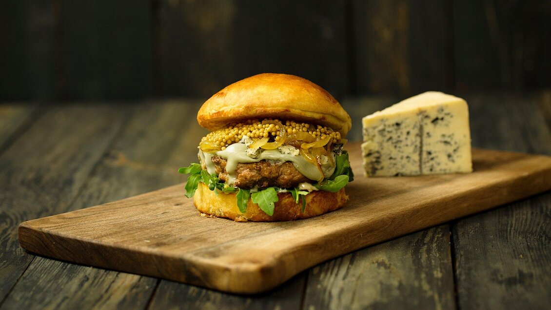 Ein saftiger Burger auf einem Holzbrett, belegt mit Salat, Zwiebeln und Senf. Neben dem Burger liegt ein Stück Käse. | © Freigeist - TG-Graphics